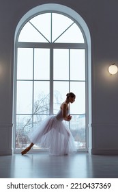 Slender Ballerina In Long White Tulle Skirt, Crouching In Bow Pose, Against Backdrop Of Huge Window