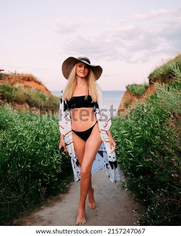 A slender beautiful blonde girl in a black swimsuit, a light pareo and a hat walks among the green hills near the ocean coast during the day.