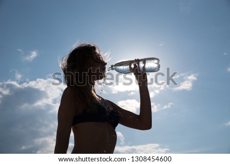 Slender beautiful girl drinks water from a bottle in a backlight against a cloudy sky.