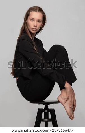 A slender beautiful teenage girl in a black jacket sat in a beautiful pose on a studio chair. Blonde fashion model girl posing. The dream is to be a model.
