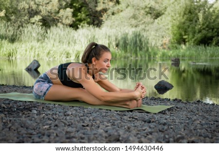 slender beautiful young girl doing yoga outdoors by the river under the bridge in the summer on the Mat