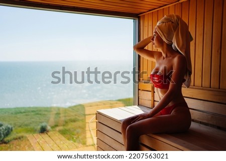 a slender and beautiful young woman in a red swimsuit sits on a bench in the sauna with the panoramic window. relax in the spa center. tourism and travel.