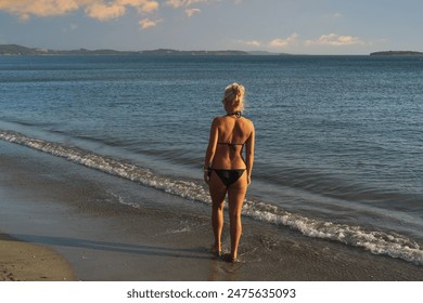 A slender blonde girl in a bikini is alone on a sandy beach in Athens, Greece on a summer day, photo at sunset from the back.  - Powered by Shutterstock