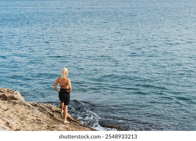 A slender blonde girl stands on the rocky shore of the Mediterranean Sea on a summer day and gets ready to swim.  - Powered by Shutterstock