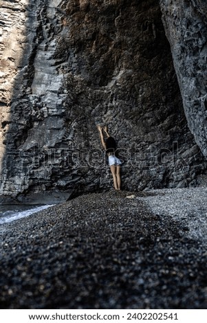 a slender brunette girl in white shorts and a black T-shirt with a beautiful figure stands near the rock of a grotto of a volcanic cave, leaning on the stones, rear view Meditation Alone with nature