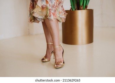 Slender Female Legs In A Summer Dress With A Floral Print And Open Summer Golden Shoes. Soft Selective Focus.