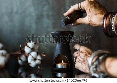 Slender female wrists painted with henna traditional Indian oriental mehndi ornaments. Hands dressed in metal bracelets and rings pour oil into aroma lamp. Branch with cotton flowers on background. 