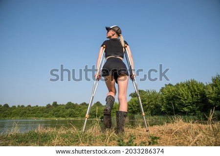 A slender girl in a baseball cap on crutches sadly looks into the distance.