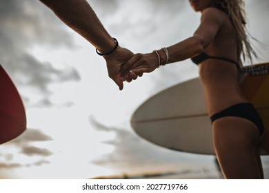 Slender Girl In Black Bathing Suit Holds Guys Hand And Carries Surfboard