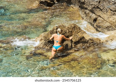 A slender girl of European appearance in a swimsuit comes out of the Mediterranean Sea from the rocky shore onto dry land.  - Powered by Shutterstock