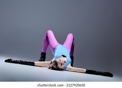 A slender girl model in bright extravagant clothes, leather gloves and high-heeled boots poses on the floor. Grey background. Black makeup and avant-garde hairstyle. Vanguard fashion. 