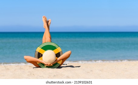 A Slender Girl On The Beach In A Straw Hat In The Colors Of The Jamaican Flag. The Concept Of A Perfect Vacation In A Resort In The Jamaica. Focus On The Hat.