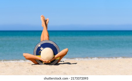 A slender girl on the beach in a straw hat in the colors of the flag of Curacao. The concept of a perfect vacation in a resort in the Curacao. Focus on the hat.