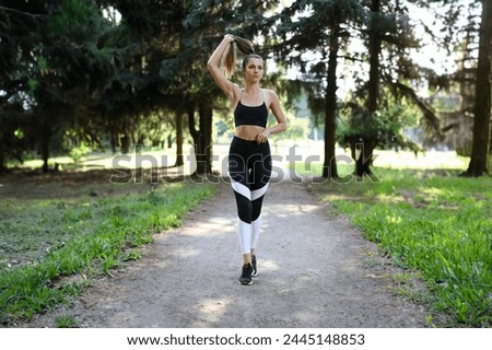 Slender girl in sportswear straightens her hair in park. Sports training at same time leads to self-discipline. Athlete circulatory system works more efficiently. Beautiful girl walks in park.