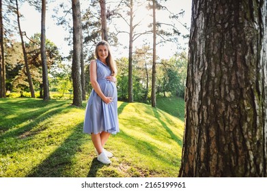 Slender happy pregnant woman in light dress posing against green trees background in park, hugging her big tummy with baby. Portrait of young expectant mother smiling outdoors. Place for text. - Shutterstock ID 2165199961