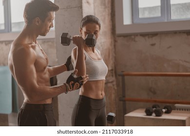 Slender lady holding dumbbells, sporty strong man helping her to correctly perform exercise for arms in the fitness club