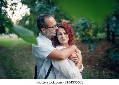 slender man with beard kisses and hugs pretty red-haired curly woman. Cute middle aged european couple hugging in the park