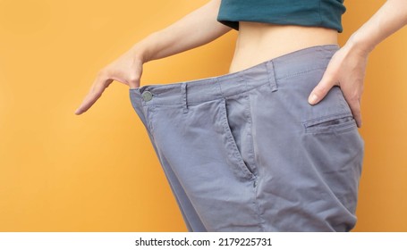 A Slender Middle-aged Woman In Large Purple Trousers Shows That They Are Too Big For Her. Orange Background, Selective Focus On The Belt Of Trousers. The Concept Of Losing Weight, Watch Your Weight.