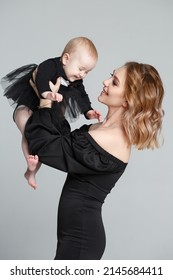 A Slender Mom Lifts The Baby Up In A Black Family Look, Isolated On A Gray Background.