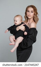 A Slender Mom Lifts The Baby Up In A Black Family Look, Isolated On A Gray Background.