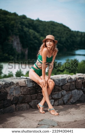 A slender positive girl with blond hair, tanned skin, dressed in a green stylish swimsuit and a hat, sits against the backdrop of a picturesque landscape with forests, a river and a waterfall.
