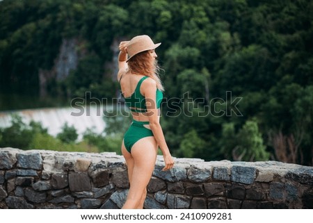 A slender positive girl with blond hair, tanned skin, dressed in a green stylish swimsuit and a hat, stands against the backdrop of a picturesque landscape with forests, a river and a waterfall.