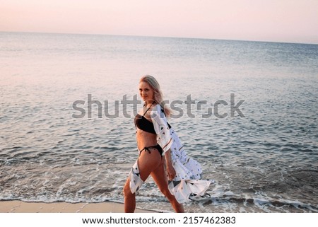 A slender smiling girl in a black bikini swimsuit with long blond hair walks along the ocean at dawn.