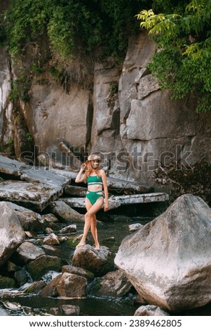 A slender smiling young girl with blond long hair, tanned skin, dressed in a green stylish swimsuit and with a small hat, stands in a river against the backdrop of picturesque high rocks.