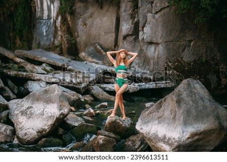 A slender smiling young girl with blond long hair, tanned skin, dressed in a green stylish swimsuit and with a small hat, stands in a river against the backdrop of picturesque high rocks.
