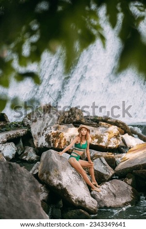 A slender sweet girl with blond hair, tanned skin, dressed in a green stylish swimsuit and with a hat, sits on stones against the backdrop of a picturesque waterfall and rests in the summer.