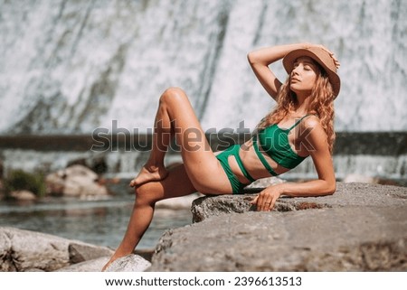 A slender sweet girl with blond hair, tanned skin, dressed in a green stylish swimsuit and with a hat, sits on stones against the backdrop of a picturesque waterfall and rests in the summer.