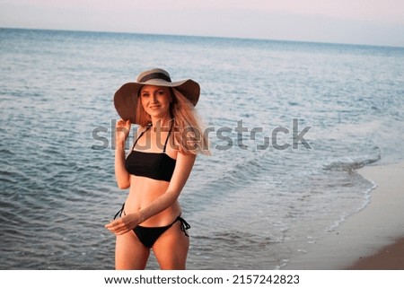 A slender tanned blonde girl with long hair in a black bikini and a hat smiles and walks along the ocean at dawn.