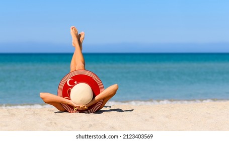 A slender tanned girl on the beach in a straw hat in the colors of the flag of Turkey. The concept of a perfect vacation in a resort in the Turkey. Focus on the hat. - Shutterstock ID 2123403569