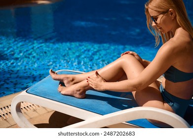 Slender woman applying sunscreen on her leg while sitting poolside. Girl applying waterproof sunscreen lotion from a bottle to her body on the pool deck. - Powered by Shutterstock