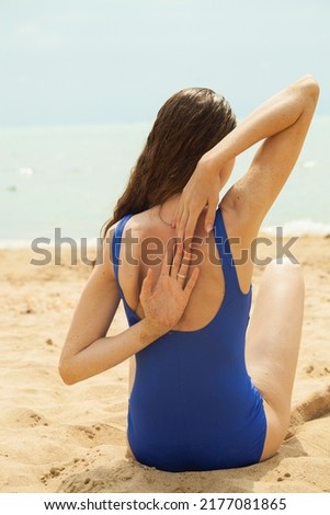a slender woman with curly hair is sitting in a blue swimsuit on a sandy beach and doing a yoga exercise sitting with her back to the camera
