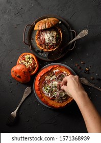 Slender Woman Hand Pouring Grated Cheese On Pumpkin Stuffed With Rice, Mushrooms And Onions And Smaller Pumpkins With Red Rice And Pearl Couscous On Black Background Top View Copy Space