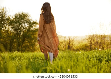 a slender woman stands in a long orange dress with her back to the camera illuminated by the sunset rays of the sun and looks towards the sky