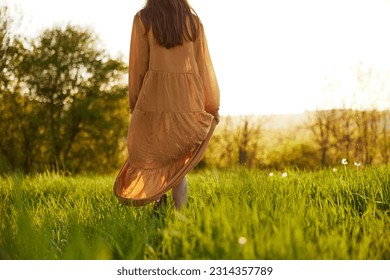 a slender woman stands in a long orange dress with her back to the camera illuminated by the sunset rays of the sun and looks towards the sky