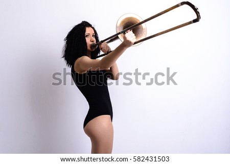 Slender young brunette girl with curly hair in a black bodysuit playing the trombone on a white background. Studio shot