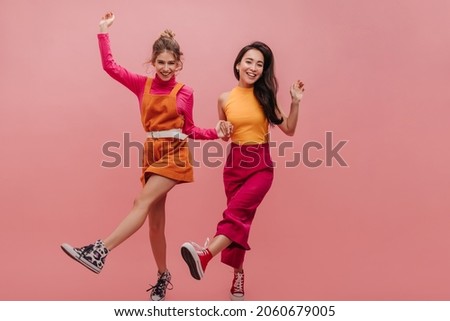 Slender young ladies from eastern countries dance holding hands on pink background. Full size image of funny girls fooling around during photo shoot. Brunette and blonde in bright casual outfits.