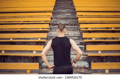 A slender young man in black sportswear stands in front of the stairs stadium stands. ready for action, step to victory, challenge. - Shutterstock ID 1384926170
