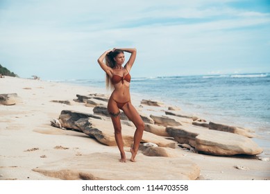 A slender young model walks along a deserted beach alone and admires the waves of the blue ocean. The girl is dressed in a trendy swimsuit, on her leg is a tattoo in the form of a garter of the bride.