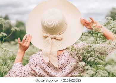 A slender young woman in a pink dress and a straw hat walking in summer meadow among wildflowers,rear view.Summer,beauty,slow life concept.