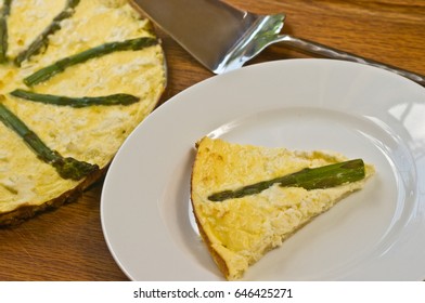 Slice of asparagus and sweet onion tart on a round, white plate with a serving utensil  and full tart all on a wood serving board paleo diet