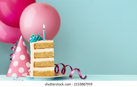 Slice of birthday cake with blue birthday candle, party hat, streamers and birthday balloons ready for a birthday party