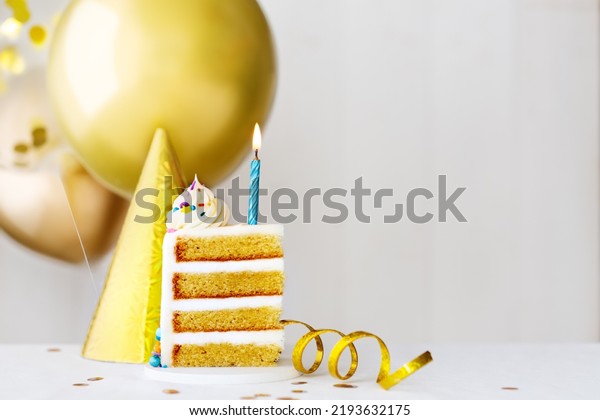 Slice of birthday cake with single blue birthday\
candle and gold birthday\
balloons