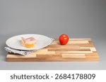 A slice of bread with ham on a plate next to a tomato on a wooden board.