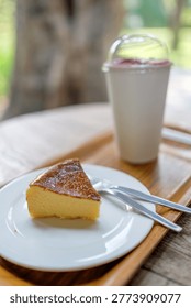 Slice of burnt basque cheesecake served on a wooden tray with a refreshing drink and a vintage style camera in a blurred cafe garden setting.