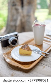 Slice of burnt basque cheesecake served on a wooden tray with a refreshing drink and a vintage style camera in a blurred cafe garden setting.