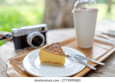 Slice of burnt basque cheesecake served on a wooden tray with a refreshing drink and a vintage style camera in a blurred cafe garden setting.